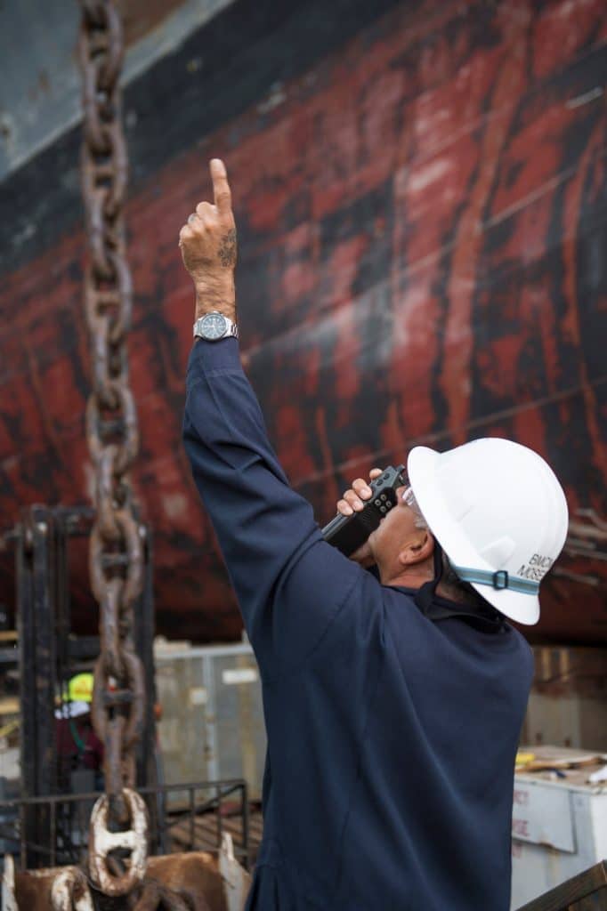 A worker in a hard hat and blue coveralls points upward while looking through binoculars near a large, weathered ship hull.