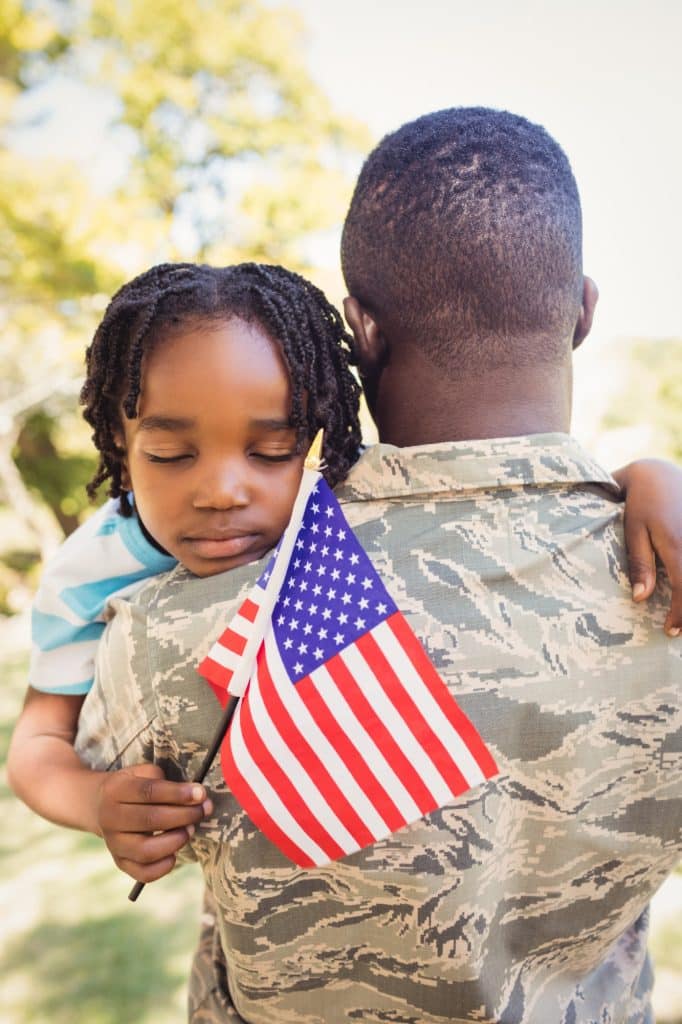 Child holding a small American flag hugs a person in military uniform outdoors, with the child's eyes closed.