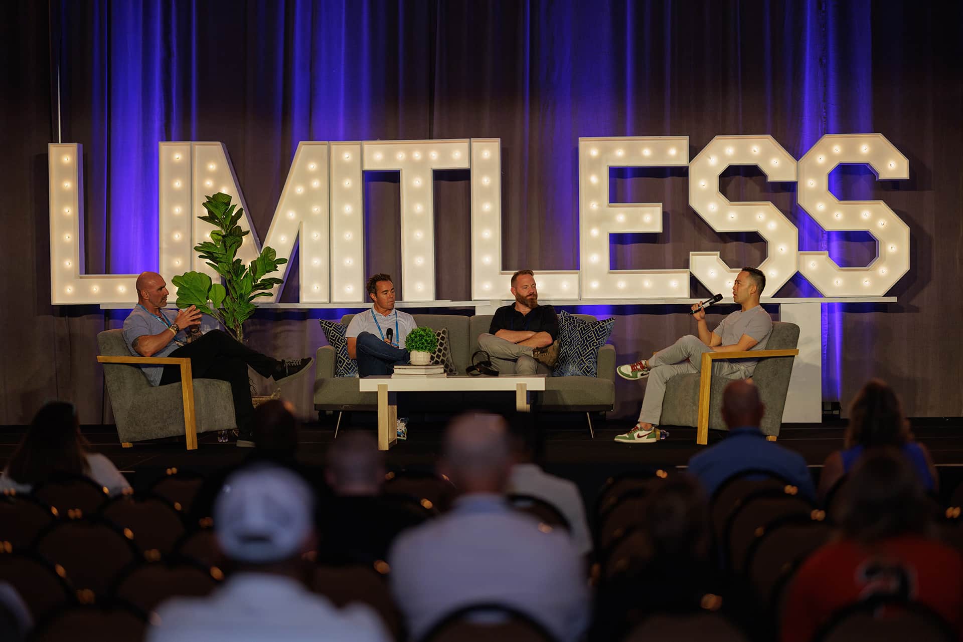 Four men sit on a stage panel in front of a large illuminated "LIMITLESS" sign, speaking to an audience in a conference setting.