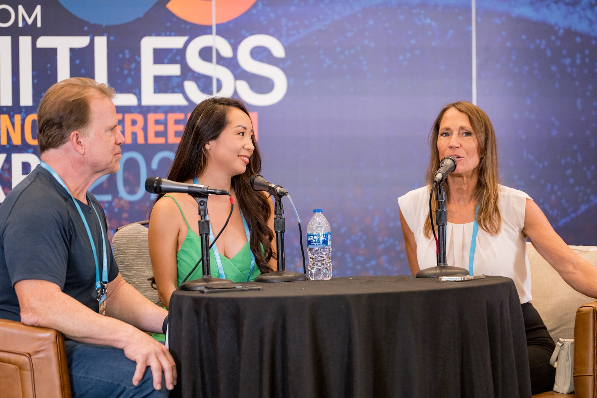 Three people sit at a round table with microphones, engaged in a panel discussion. A water bottle is on the table and an event banner is visible in the background.