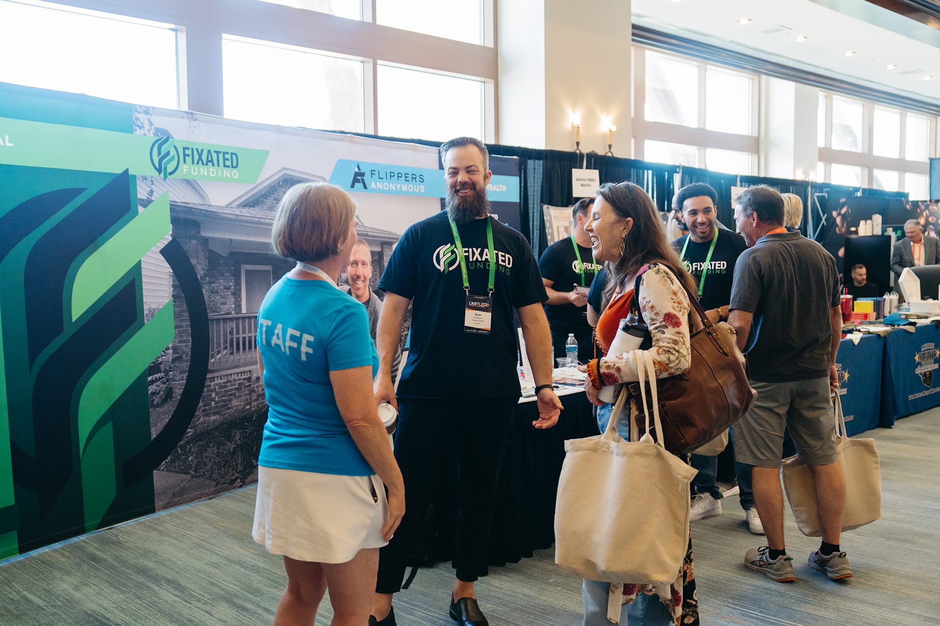 People interact and smile at an event booth for Fixated Funding. Some wear branded shirts, and informational materials are visible on tables.