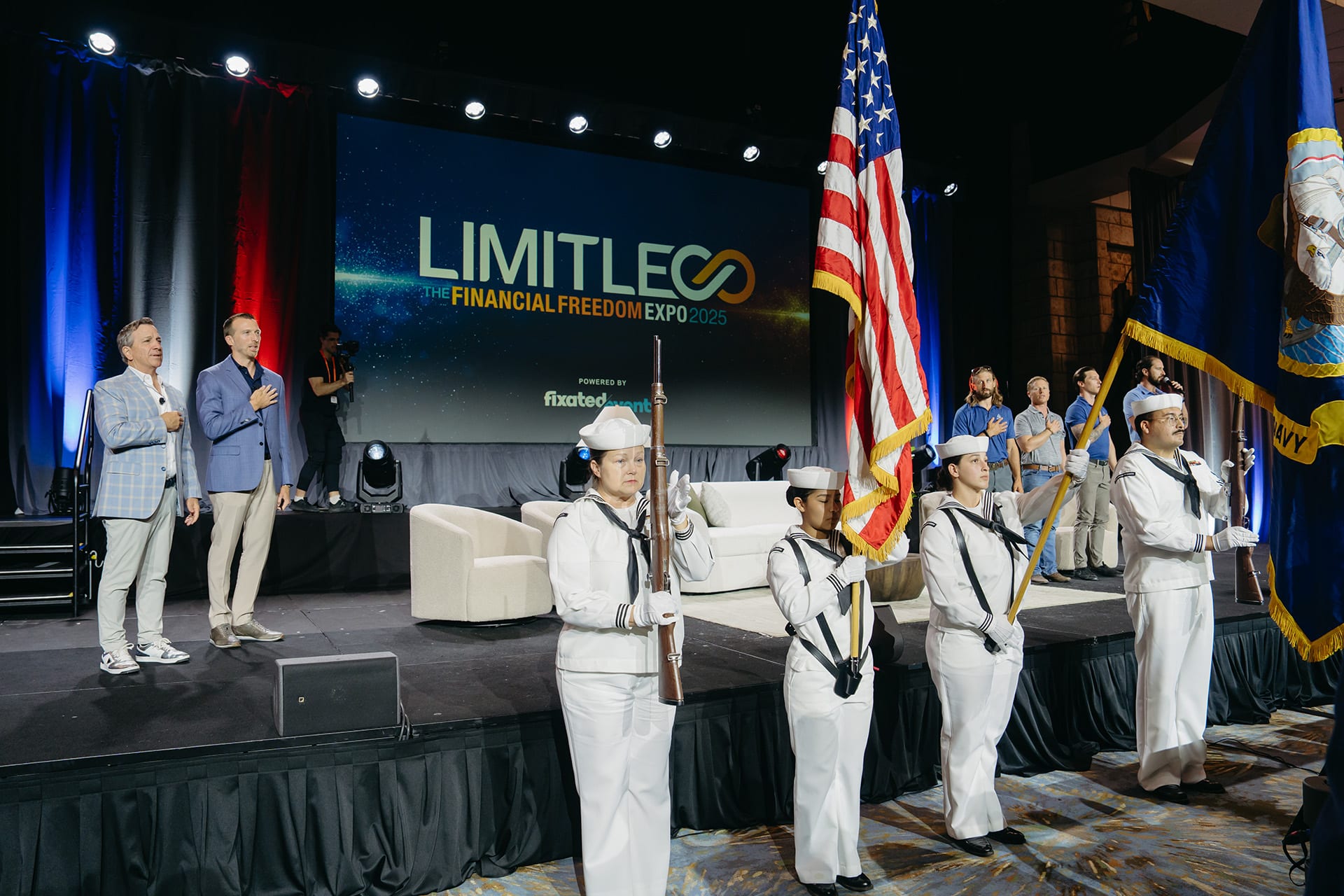 A color guard presents flags on stage while two men stand with hands over hearts at the LIMITLESS Financial Freedom Expo 2023 event.