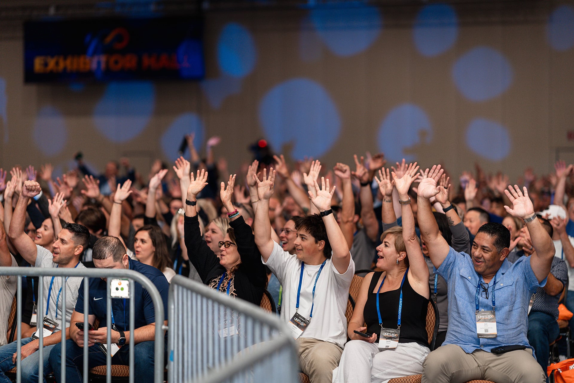 A large audience seated in rows raises their hands enthusiastically at an indoor event, with an "Exhibitor Hall" sign visible in the background.