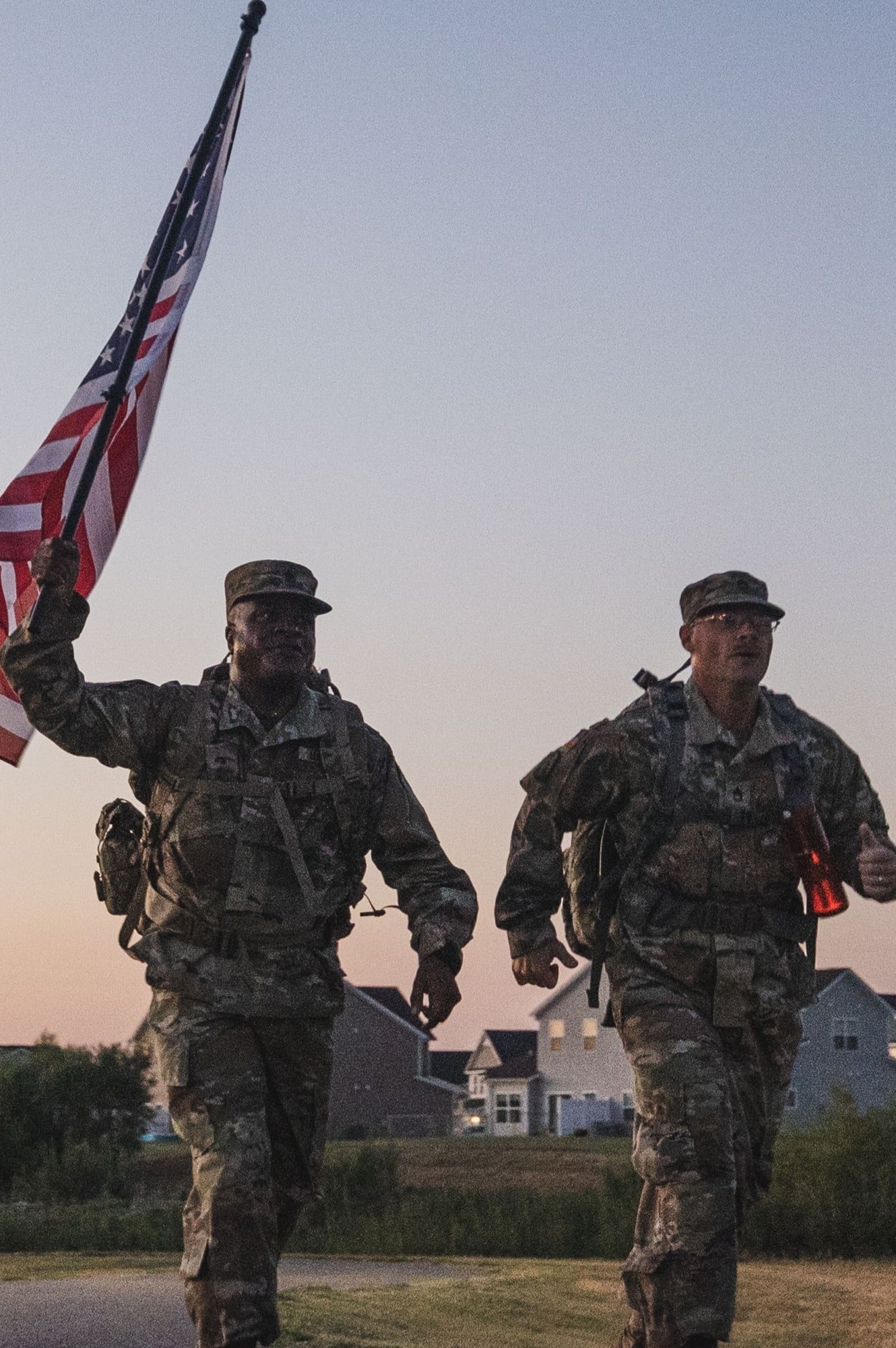 Two soldiers in camouflage uniforms run outdoors at dusk; one carries an American flag. Houses and trees are visible in the background.