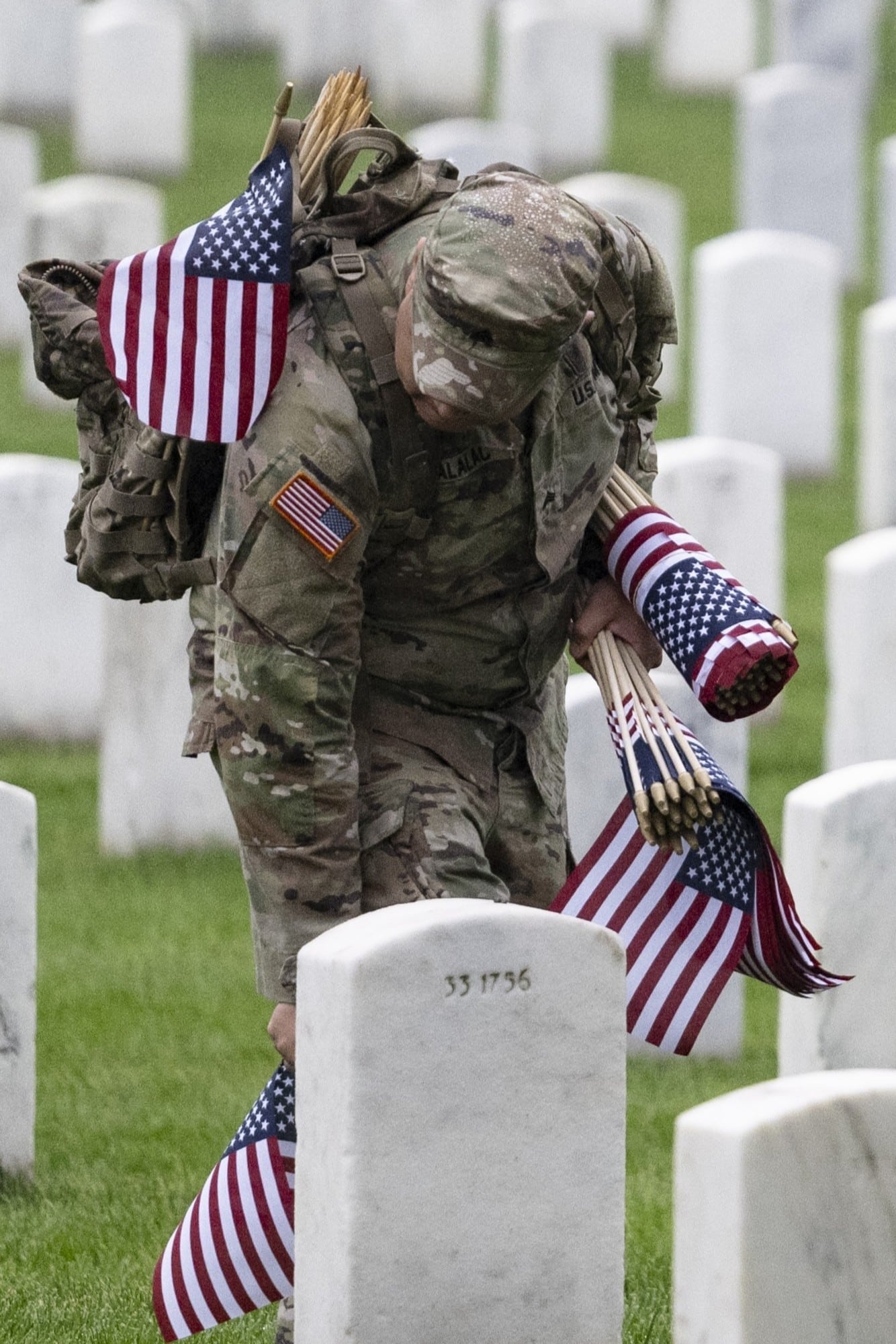 A U.S. soldier places small American flags at graves in a cemetery, carrying more flags under one arm and wearing military uniform.