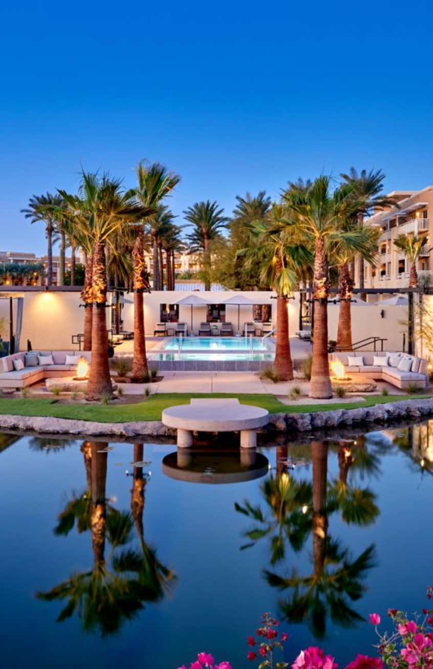 A modern resort pool area with palm trees, lounge seating, and a central pond reflecting the scene at sunset.