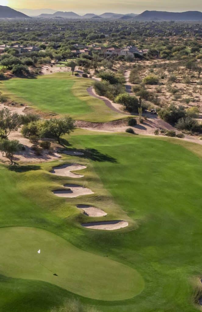 Aerial view of a desert golf course with green fairways, sand bunkers, scattered trees, and nearby houses, surrounded by arid landscape and distant mountains.