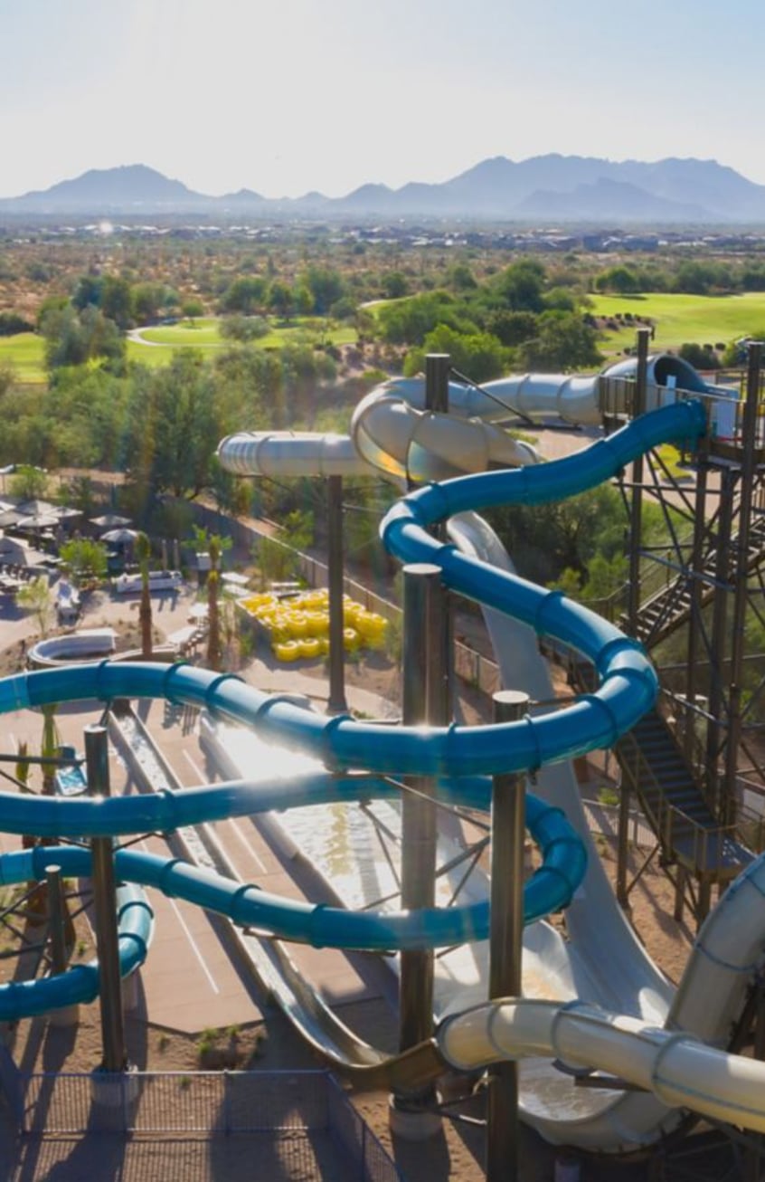 A view of winding blue and white water slides at a water park, with green fields, trees, and mountains visible in the background.