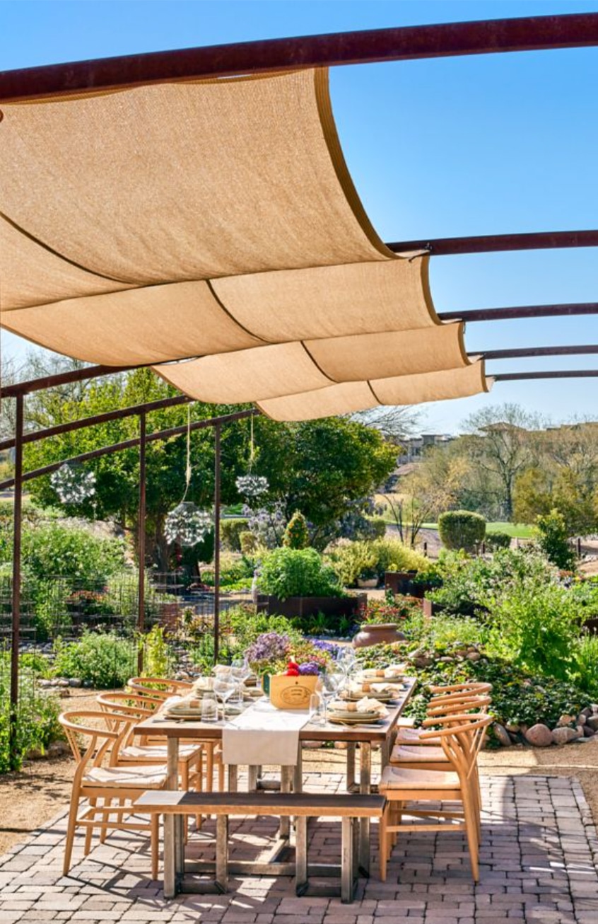 A dining table set for a meal sits under a fabric canopy in a garden, surrounded by greenery and wooden chairs on a paved patio.
