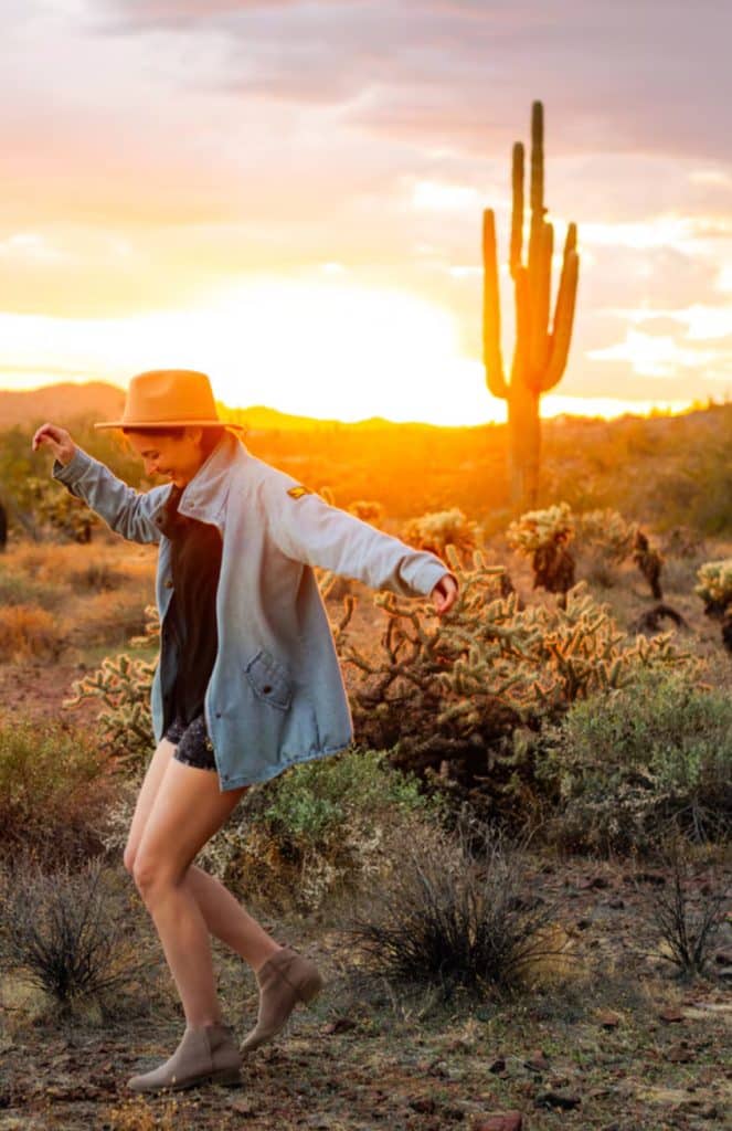 Person in a hat and jacket walking playfully in a desert landscape at sunset with a large cactus in the background.
