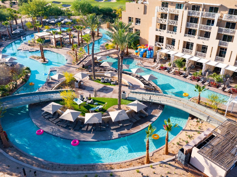 Aerial view of a hotel with a large outdoor pool, lazy river, lounge chairs, umbrellas, palm trees, and colorful inner tubes.