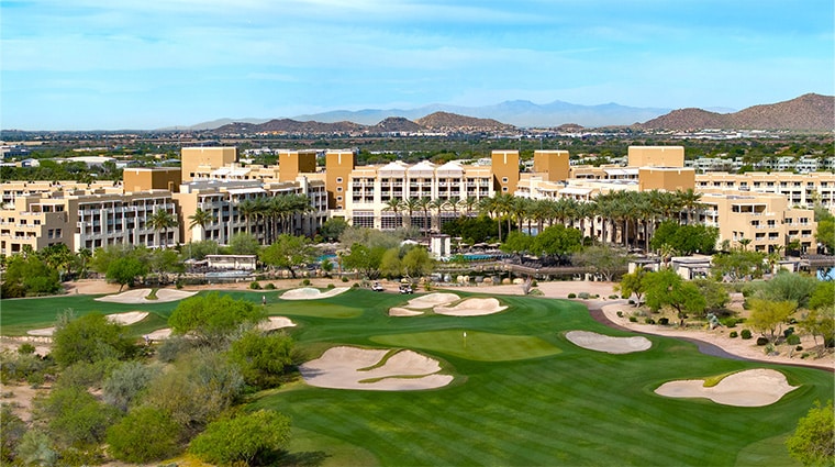 A large resort hotel is situated behind a golf course with sand bunkers and green fairways, surrounded by desert landscape and distant mountains.