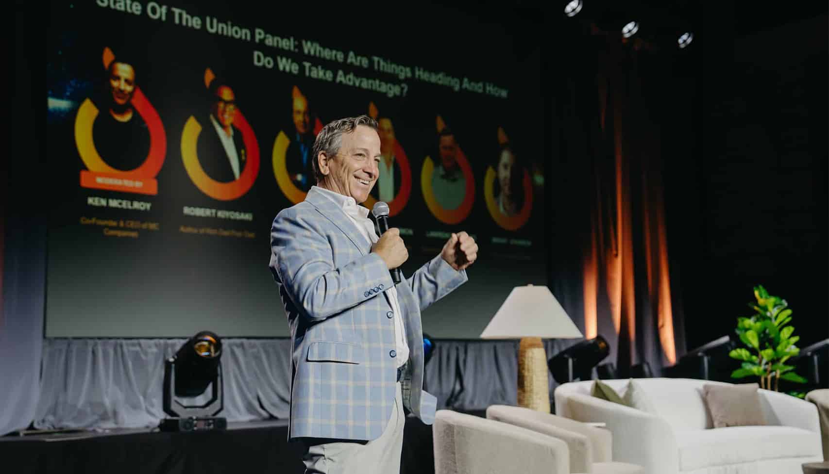 A man in a light plaid blazer speaks on stage with a microphone. Behind him is a large screen displaying a panel discussion topic and panelist names.