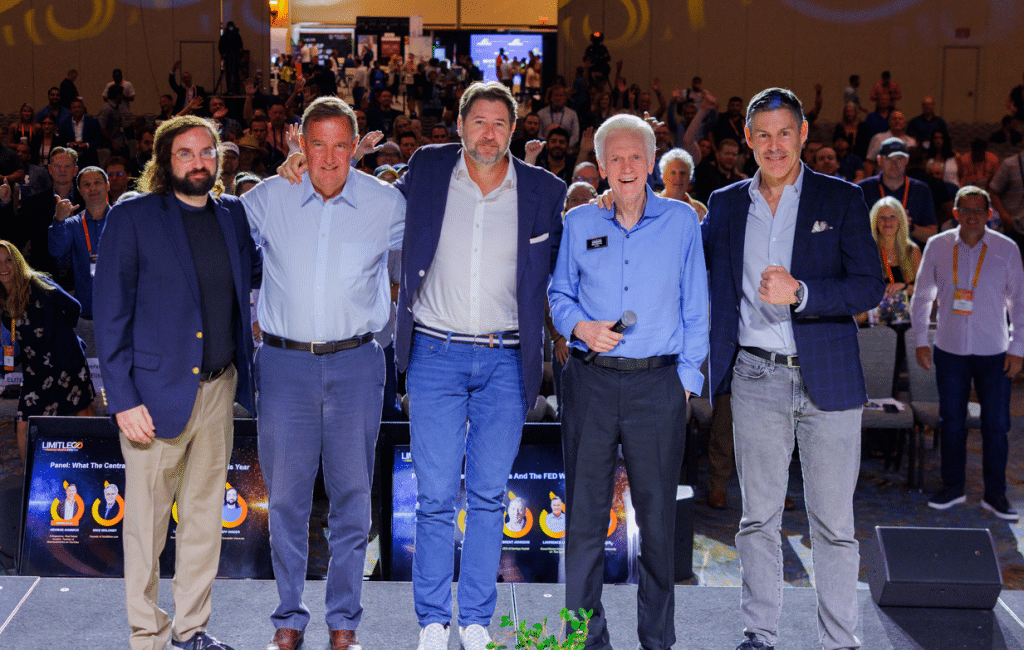 Five men stand together on a stage facing the camera, with an audience seated and standing in the background at an indoor event or conference.