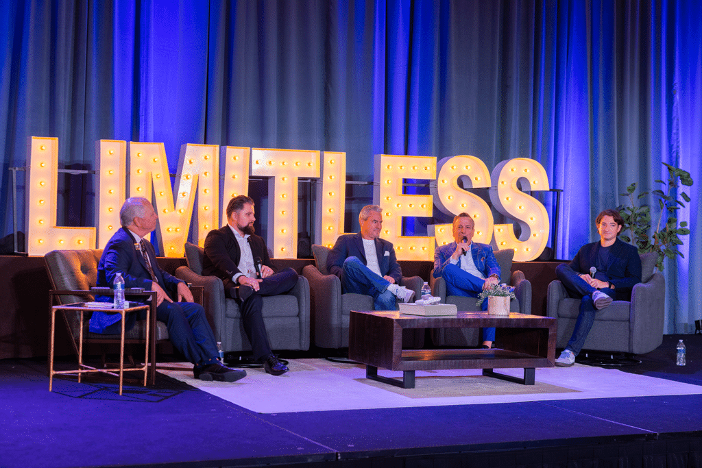 Five men in suits sit on a stage panel in front of large illuminated letters spelling "LIMITLESS," with a blue curtain backdrop and a coffee table in front of them.