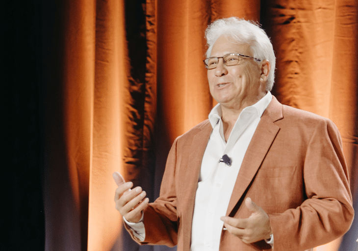 An older man in a rust-colored blazer and glasses speaks on stage in front of orange curtains, with a microphone clipped to his shirt.