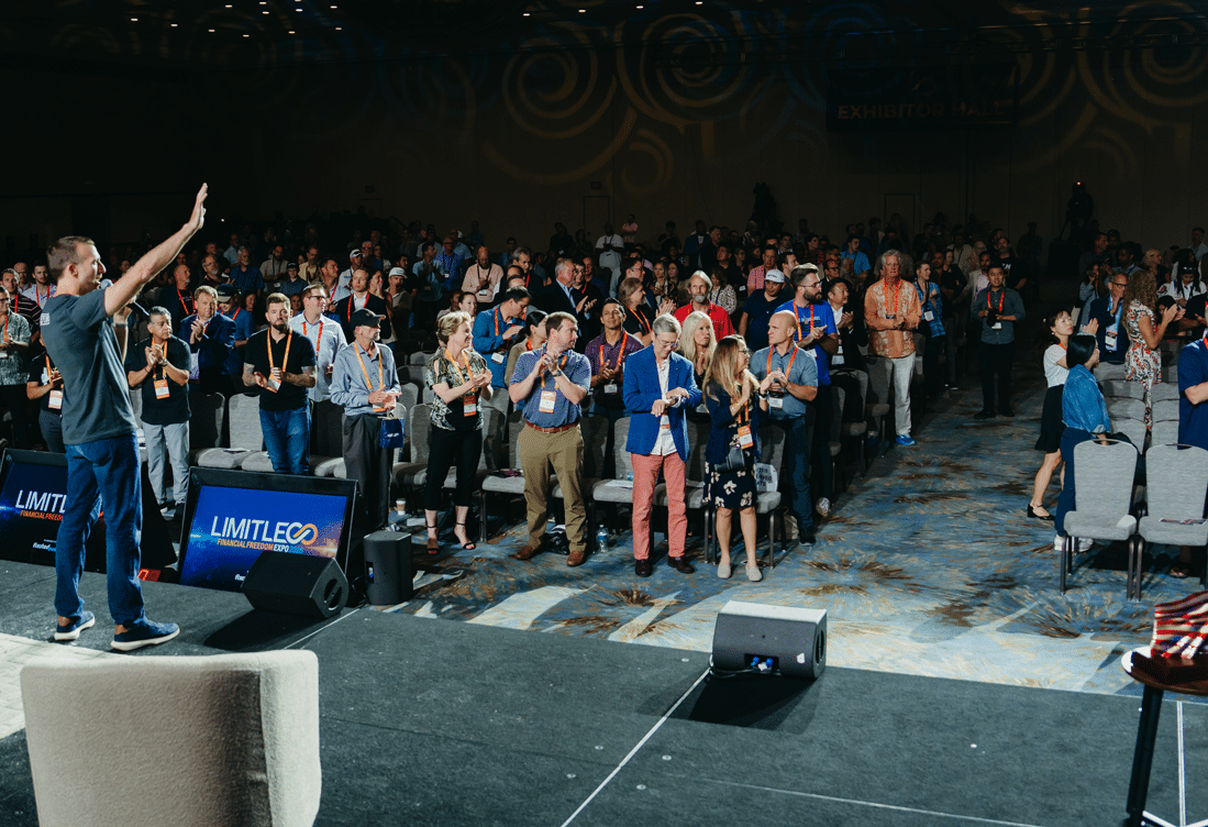 A speaker on stage addresses a large, standing audience at a conference. Many attendees are clapping, with name badges visible and large screens reading "LIMITLESS" in the foreground.