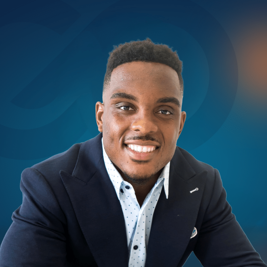 A man in a navy suit and white polka dot shirt smiles at the camera against a blue gradient background.