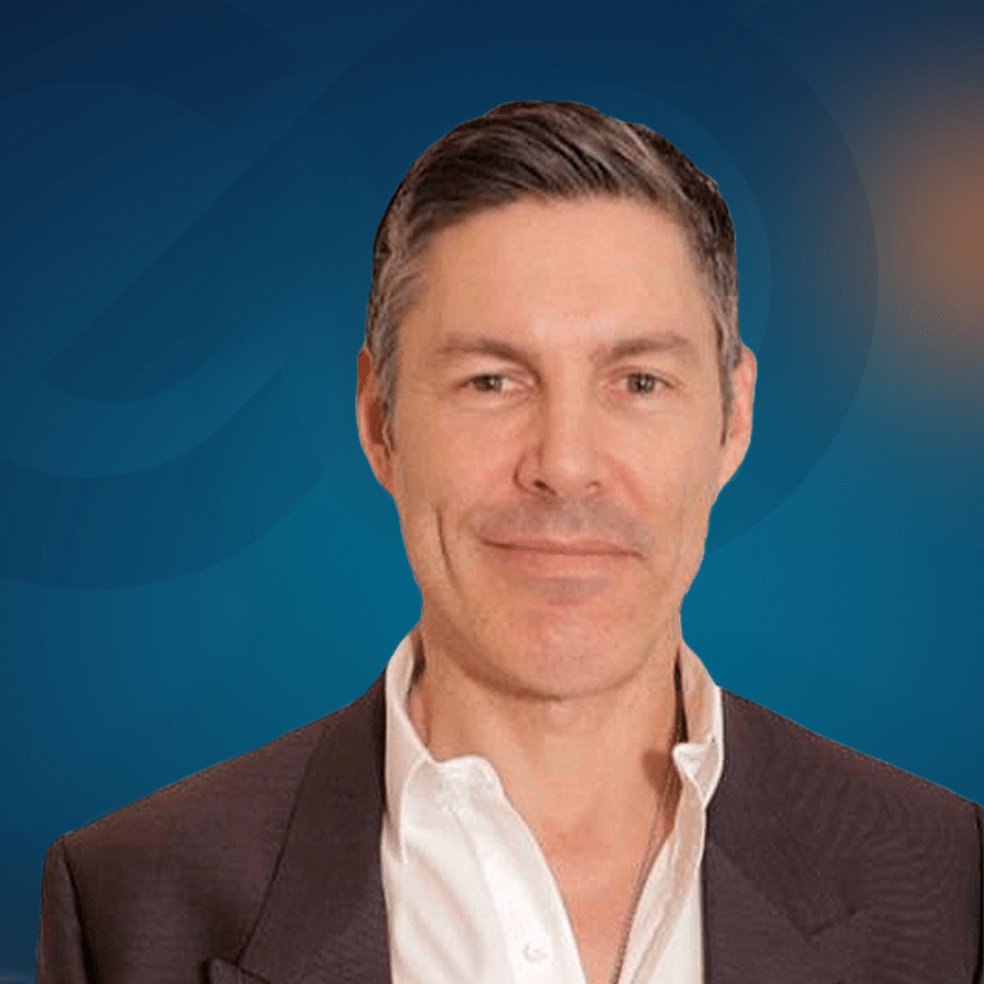 A man with short dark hair wearing a brown jacket and open-collared white shirt poses in front of a blue background with a faint circular design.