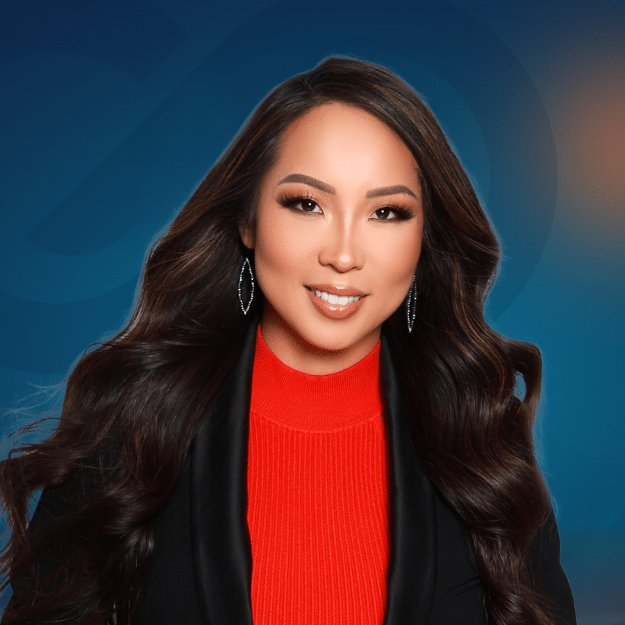 Woman with long wavy dark hair, wearing a red turtleneck and black blazer, smiling in front of a blue gradient background.