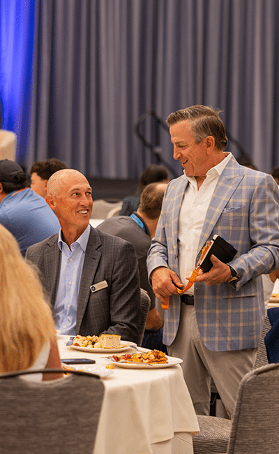 Two men in suits talking and smiling at a banquet table with food, in a conference or event setting with other attendees in the background.