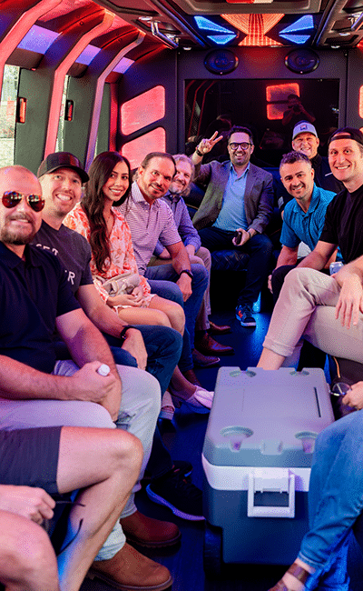 A group of people sits and smiles inside a party bus with colorful lights, some facing the camera, one person in back making a peace sign.