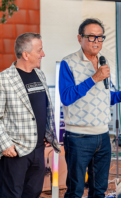 Two men stand together at an event; one holds a microphone while speaking, and the other, smiling, looks at him.