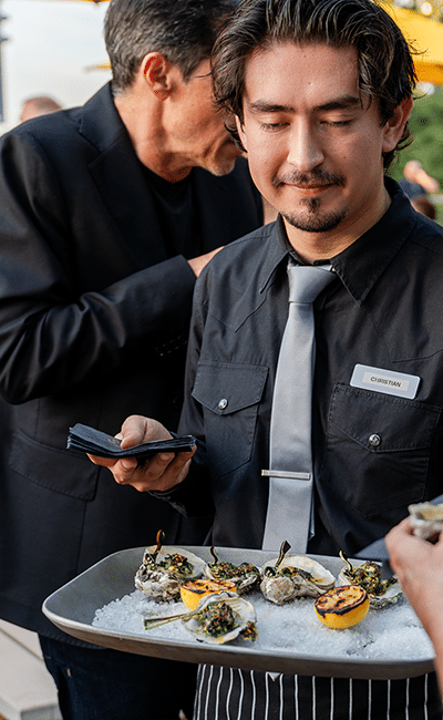 A server in a black shirt and gray tie holds a tray of oysters on ice with grilled lemon halves at an event.