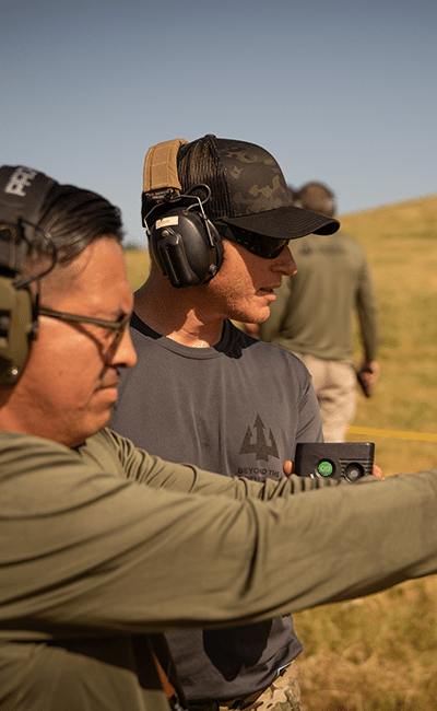 Two men wearing ear protection and hats stand outdoors on a grassy field, one pointing ahead while the other holds a small device.