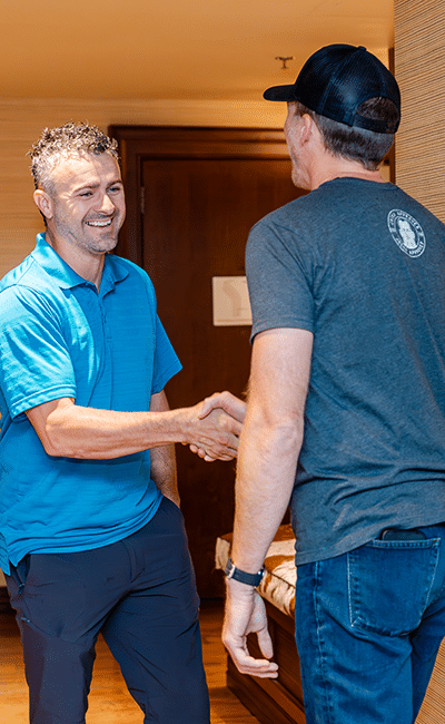 Two men are standing indoors, facing each other and shaking hands. One man is wearing a blue polo shirt; the other is in a grey t-shirt and black cap.